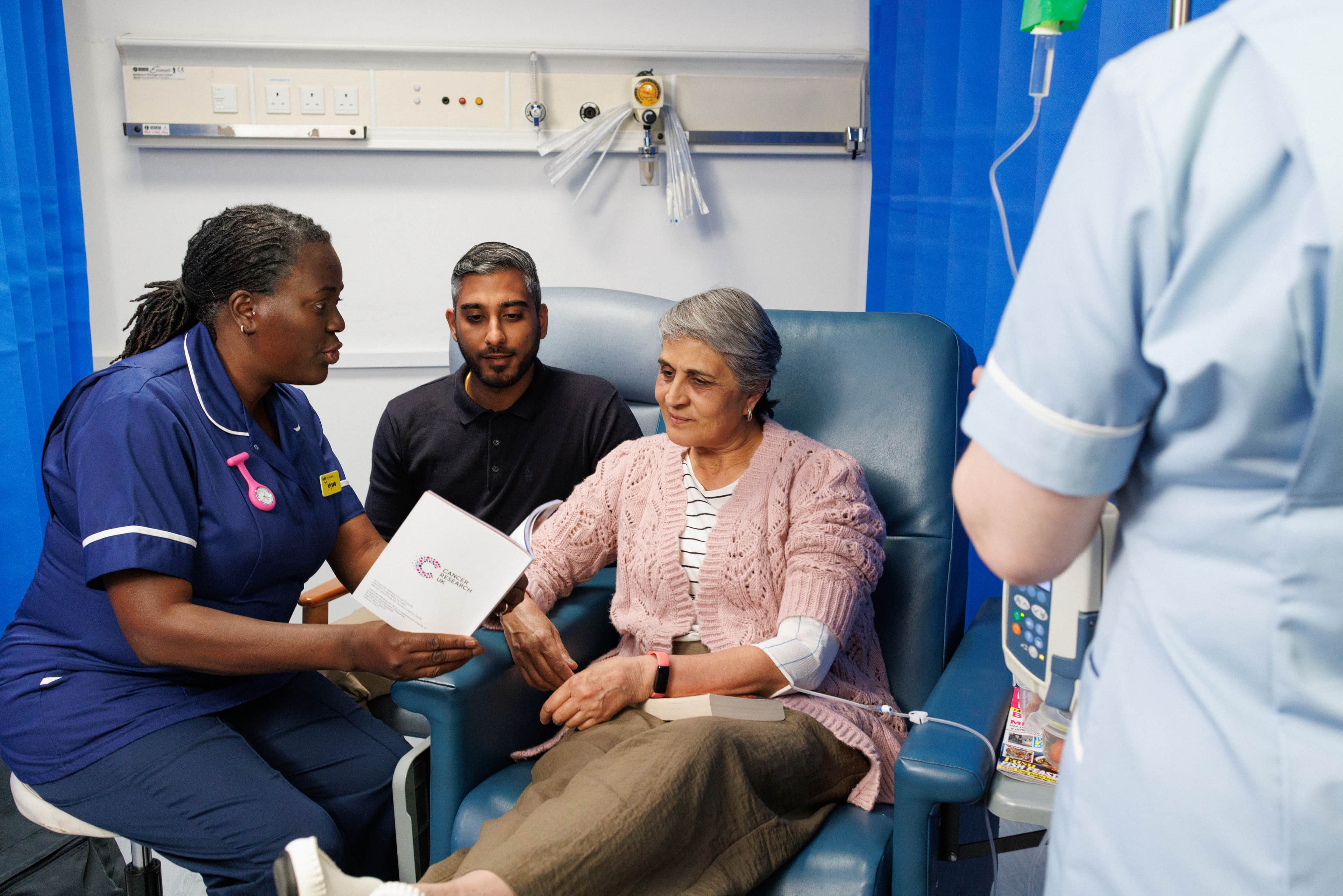 Patient sat with a family member and a health professional while having chemo. Nurse shows them a Cancer Research UK leaflet.