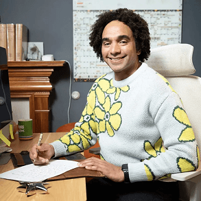 An image of Joseph Coelho smiling sitting at a desk while writing on a piece of paper.