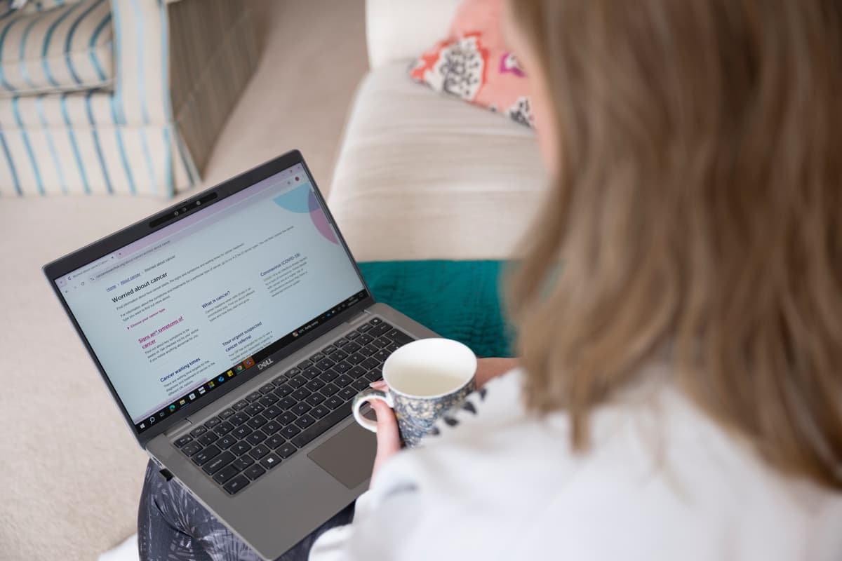 Person holding a mug looks at a laptop showing a webpage about cancer.