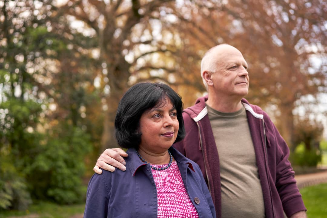 Older couple standing outdoors, man's arm around woman.