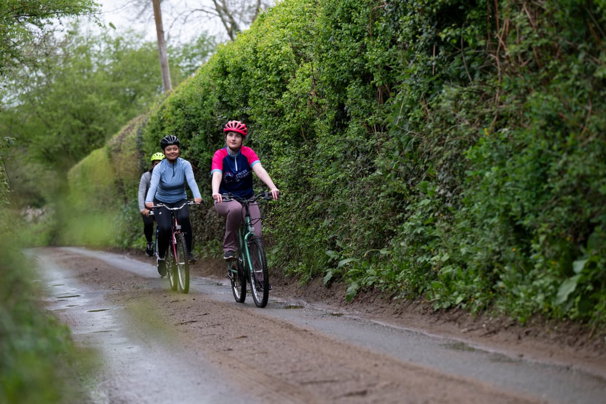 Two cyclists riding along a narrow, tree-lined country road on a sunny day.
