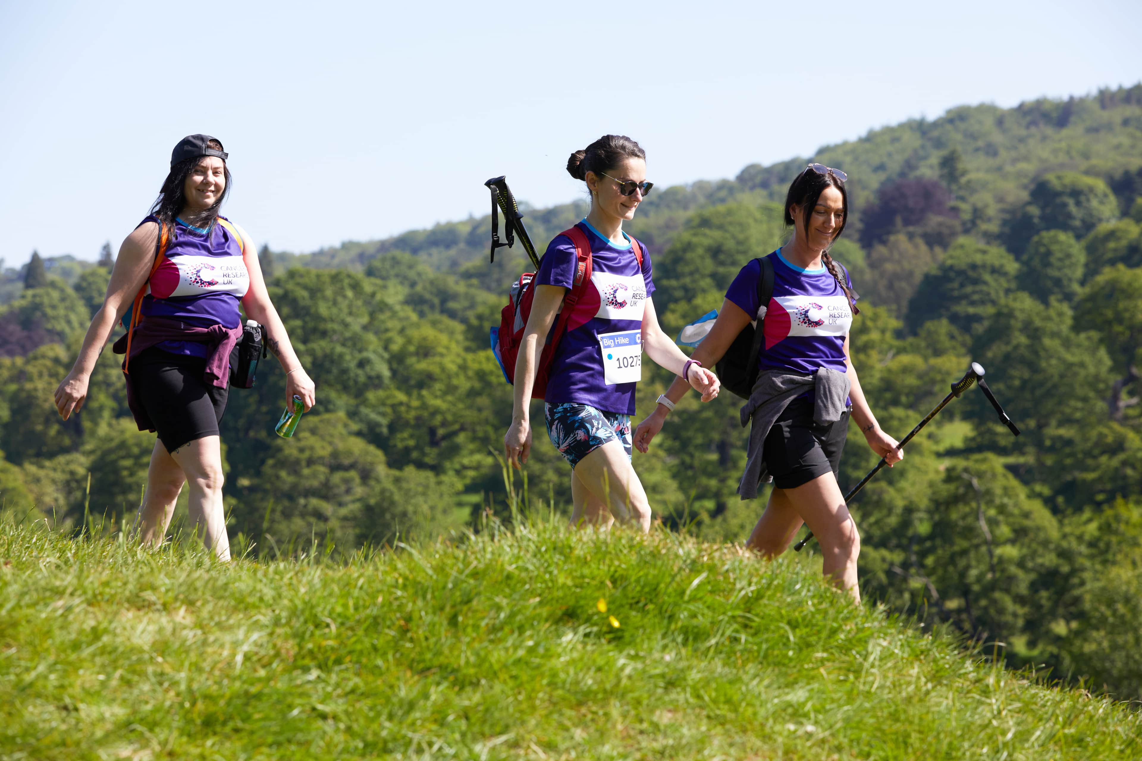 Three Big Hike Participants walk across a grassy hill, carrying backpacks, with a green, forested hillside in the background.