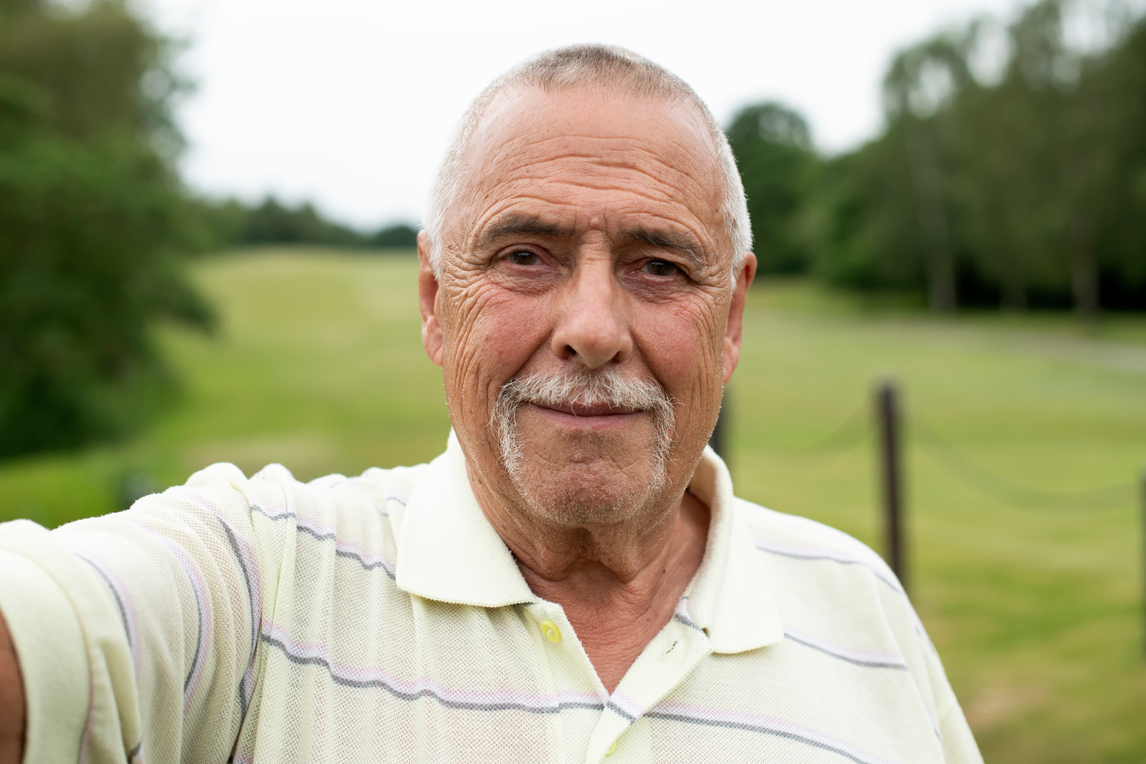 Picture of bowel cancer patient Alan, wearing a yellow polo shirt stood in a green field.