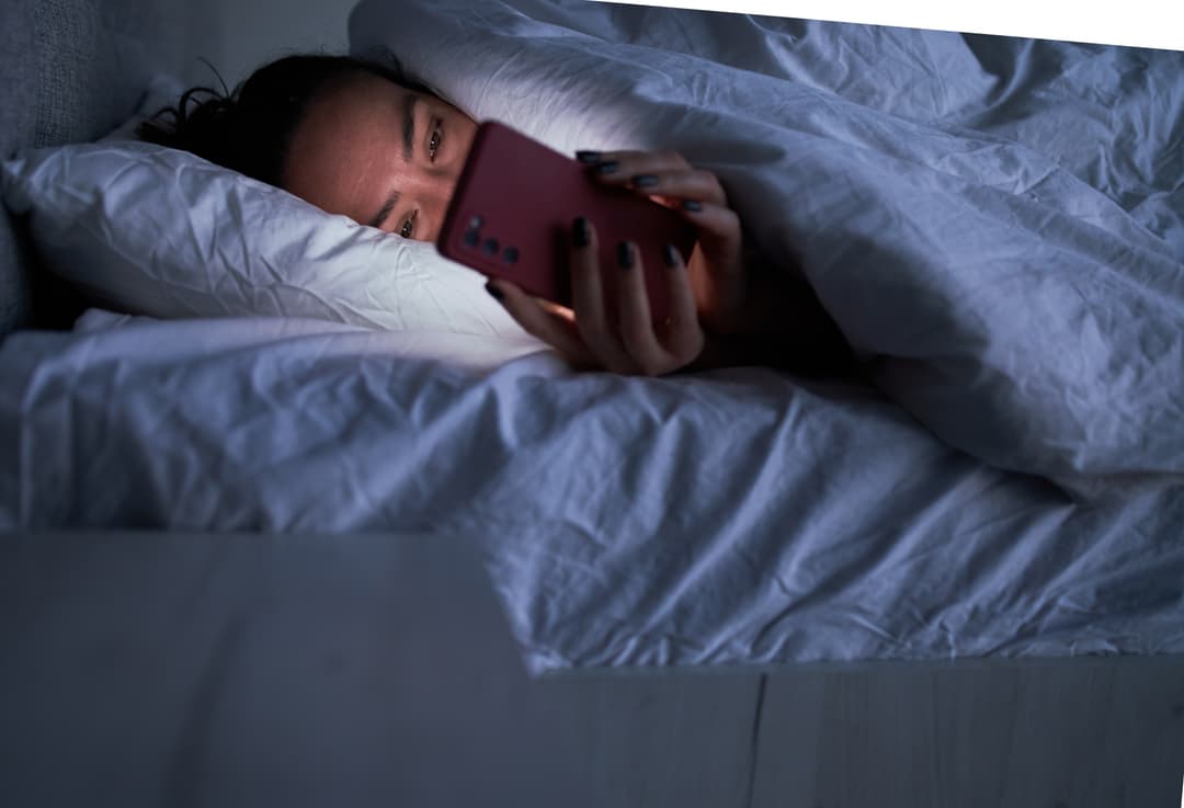 Unrecognizable young female lying in soft bed under warm duvet and using smartphone during insomnia at night.