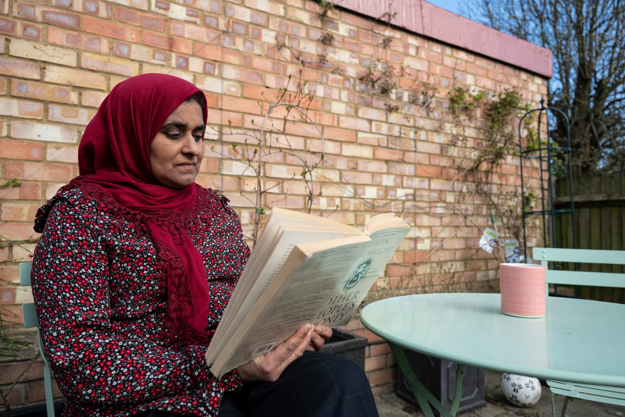 Photograph showing a woman with a head scarf reading a book in the garden.
