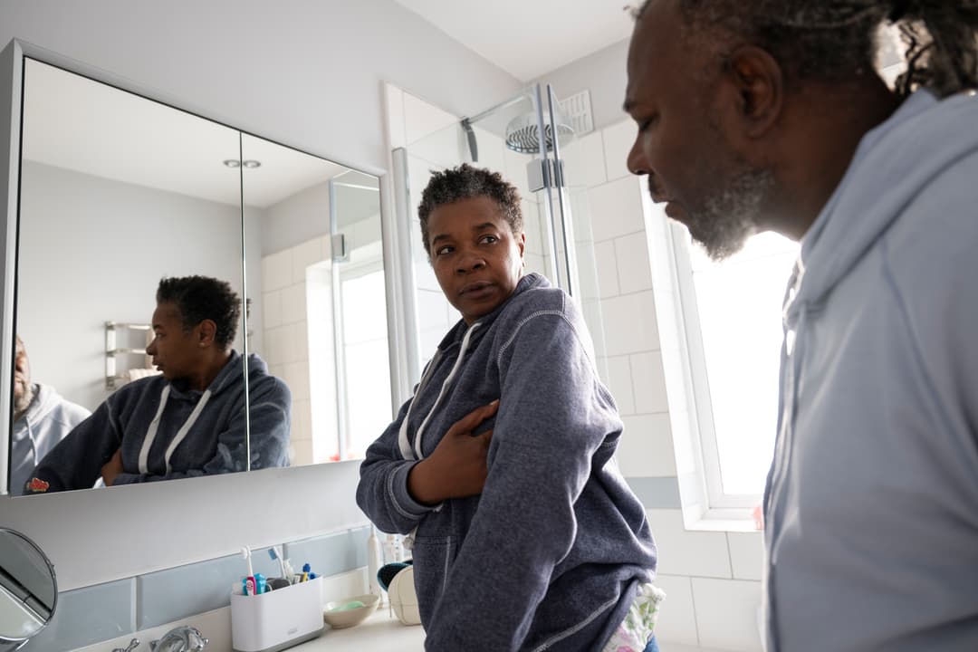 An adult looking concerned in a bathroom with another adult, with her reflection visible in the mirror.