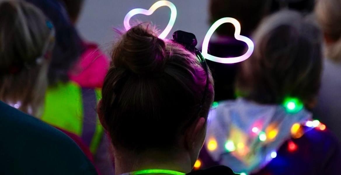 Person wearing glowing heart-shaped headband and colourful lights at a night event.