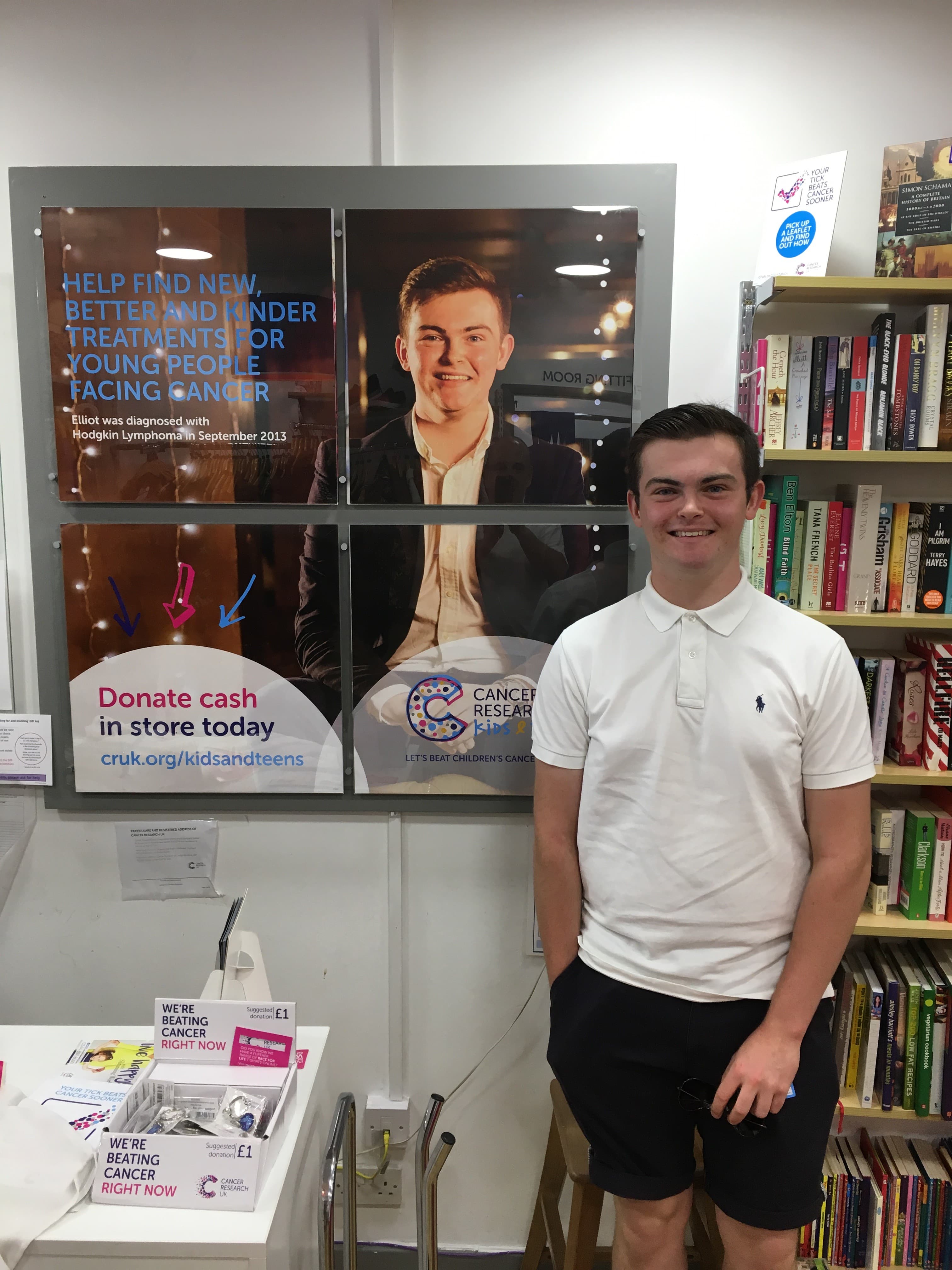 Elliot, a Hodgkin lymphoma patient, standing in a Cancer Research UK charity shop next to a poster of himself, smiling.