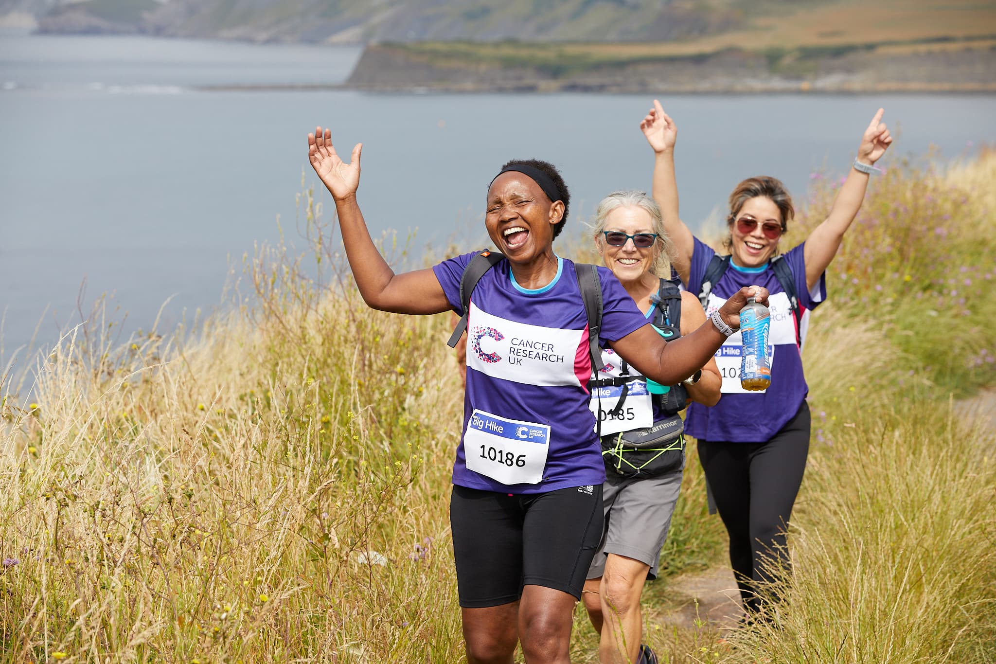 Three excited Big Hike participants are cheering with their arms up in the air on a grassy hill overlooking a body of water.