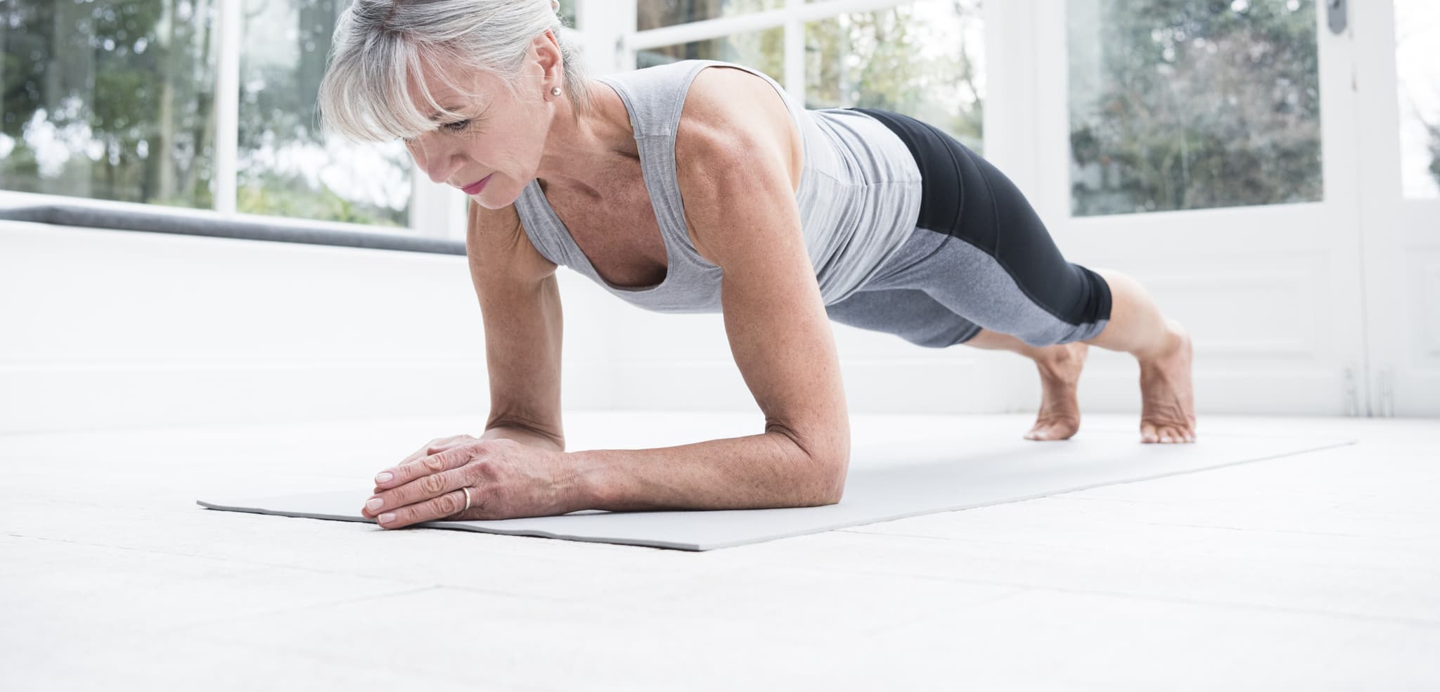 A photo of an older woman wearing gym clothes completing a plank in a bright room indoors.