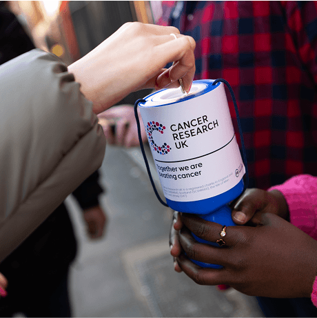 A photo of a a hand putting money into a Cancer Research UK collection pot.