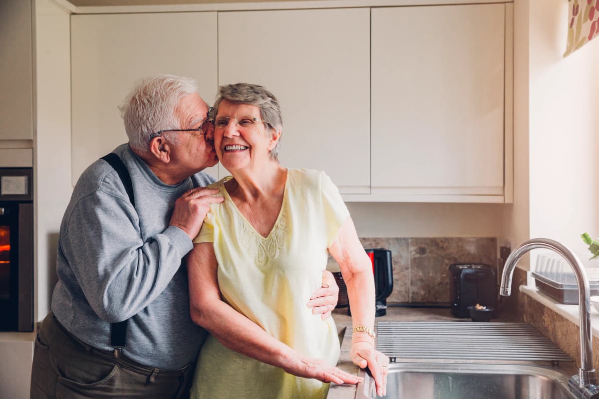 An old couple hugging each other in the kitchen and smiling.