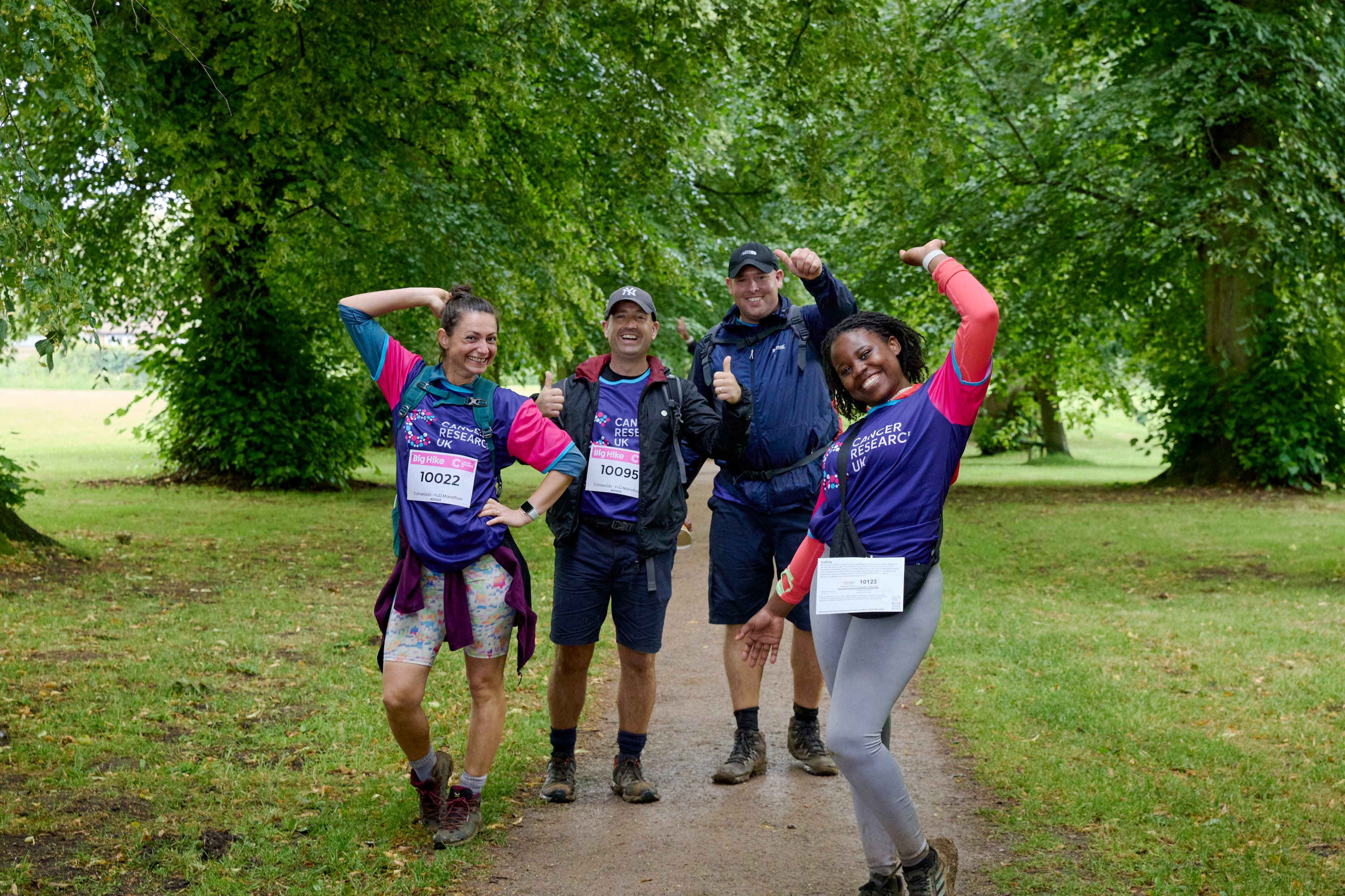 Four excited Big Hike participants are posing with their arms up on a dirt path lined with trees.