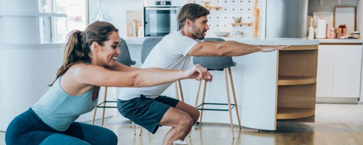 Side-shot of a man and woman side by side in a kitchen doing squats.