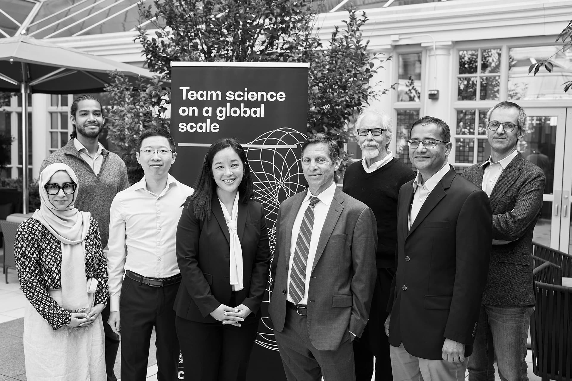Eight members of Team eDyNAmiC stand together smiling, in front of a banner that says 'Team science on a global scale'.