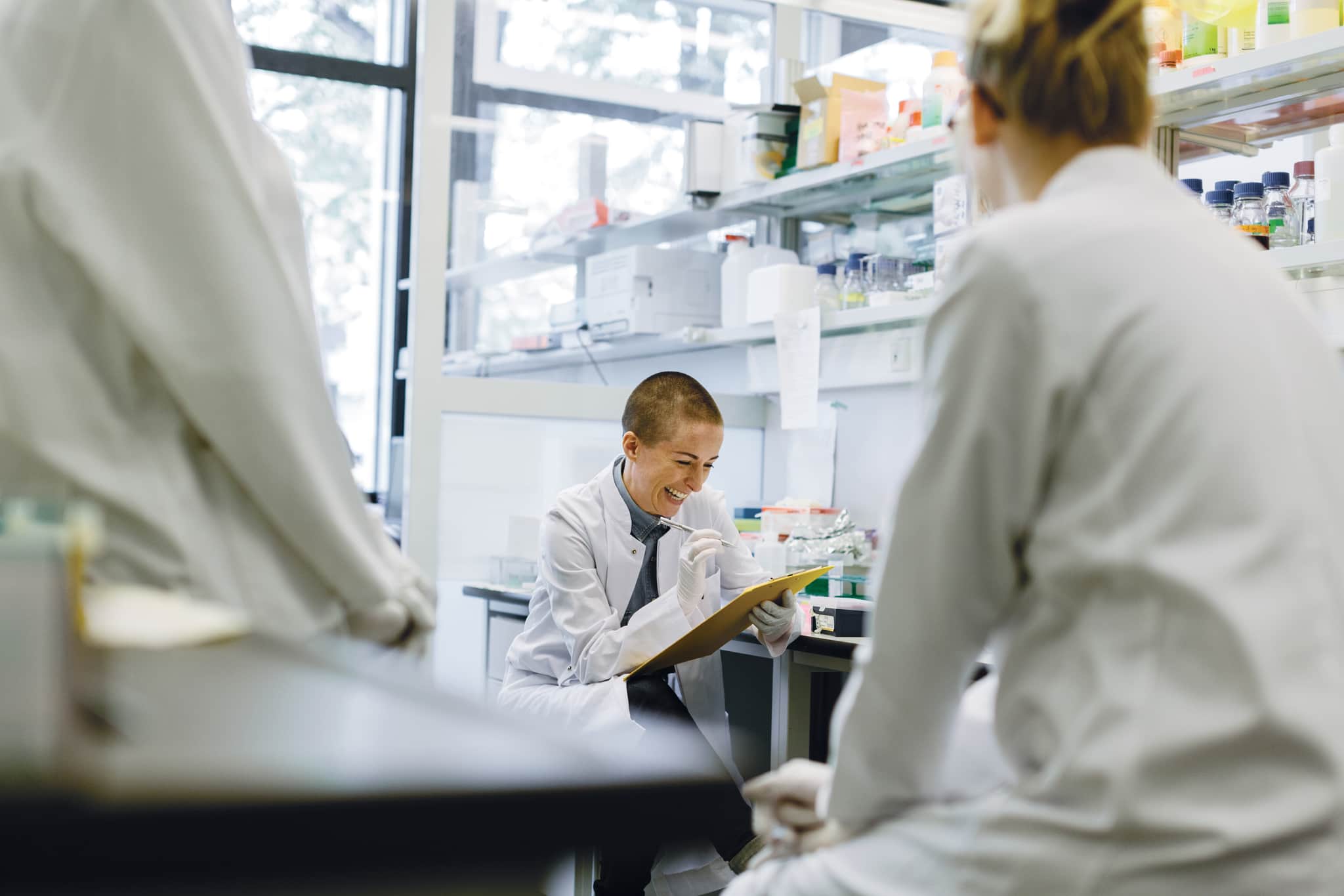 Three scientists are in a laboratory discussing their work. A scientist is holding a clipboard and writes down some data.