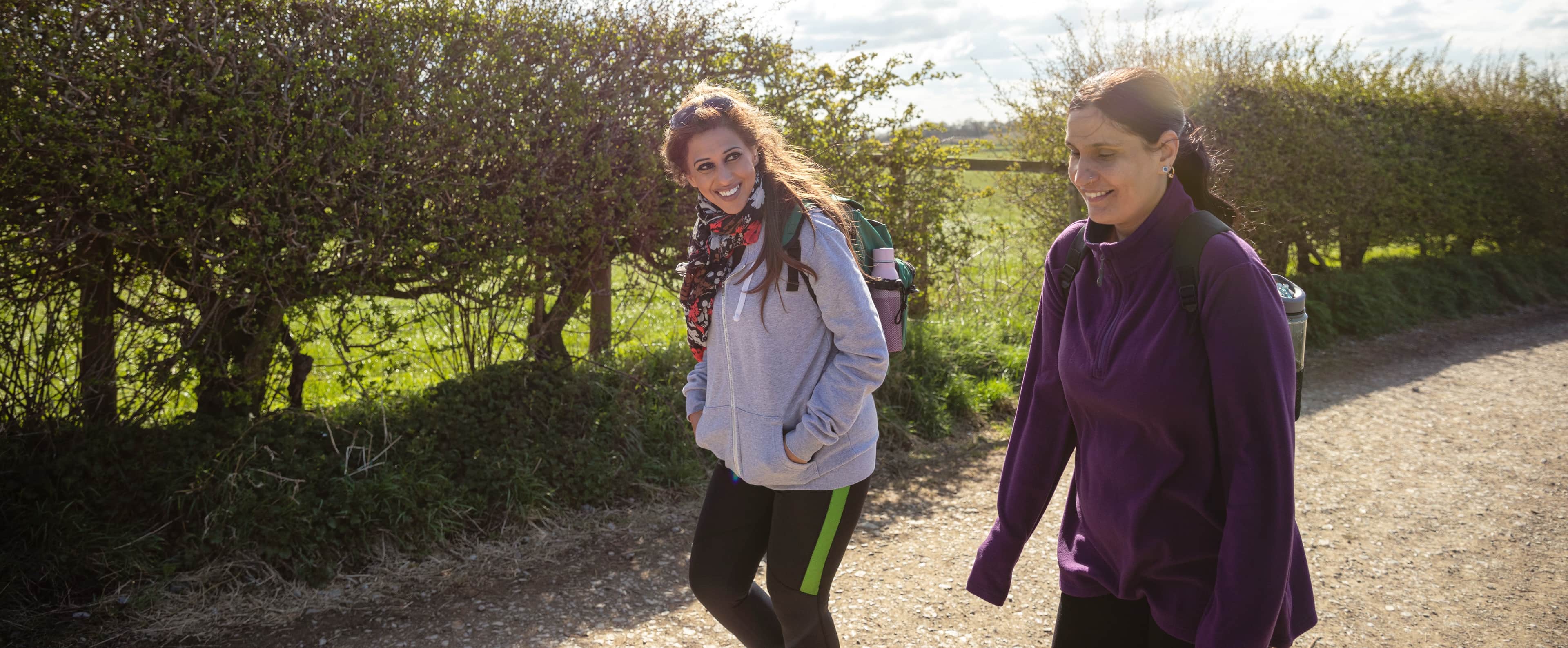 Two women walking along a sunny countryside path beside tall hedgerows.