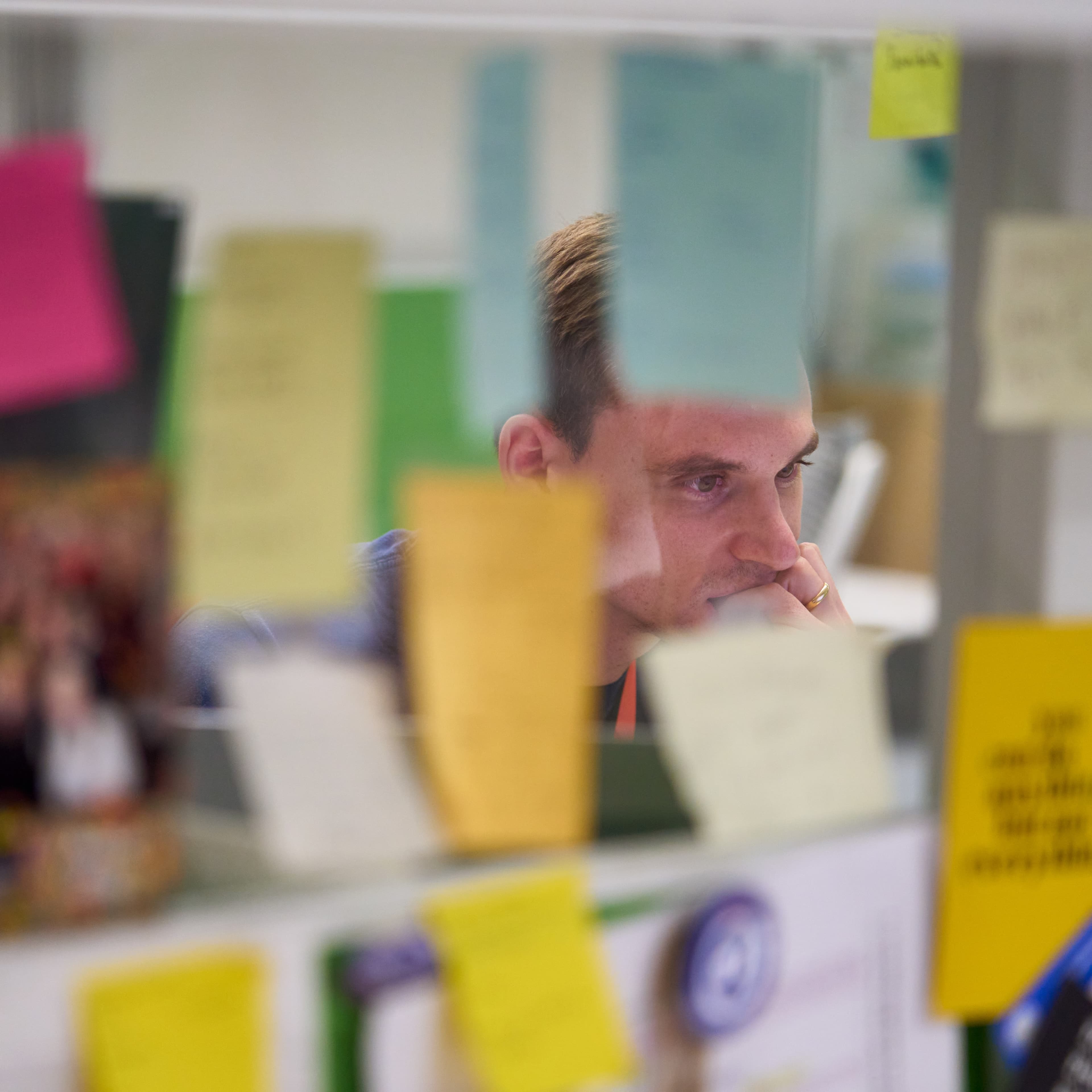 Shows a man deep in thought at a desk. In the foreground is a clear board with many sticky notes on it.