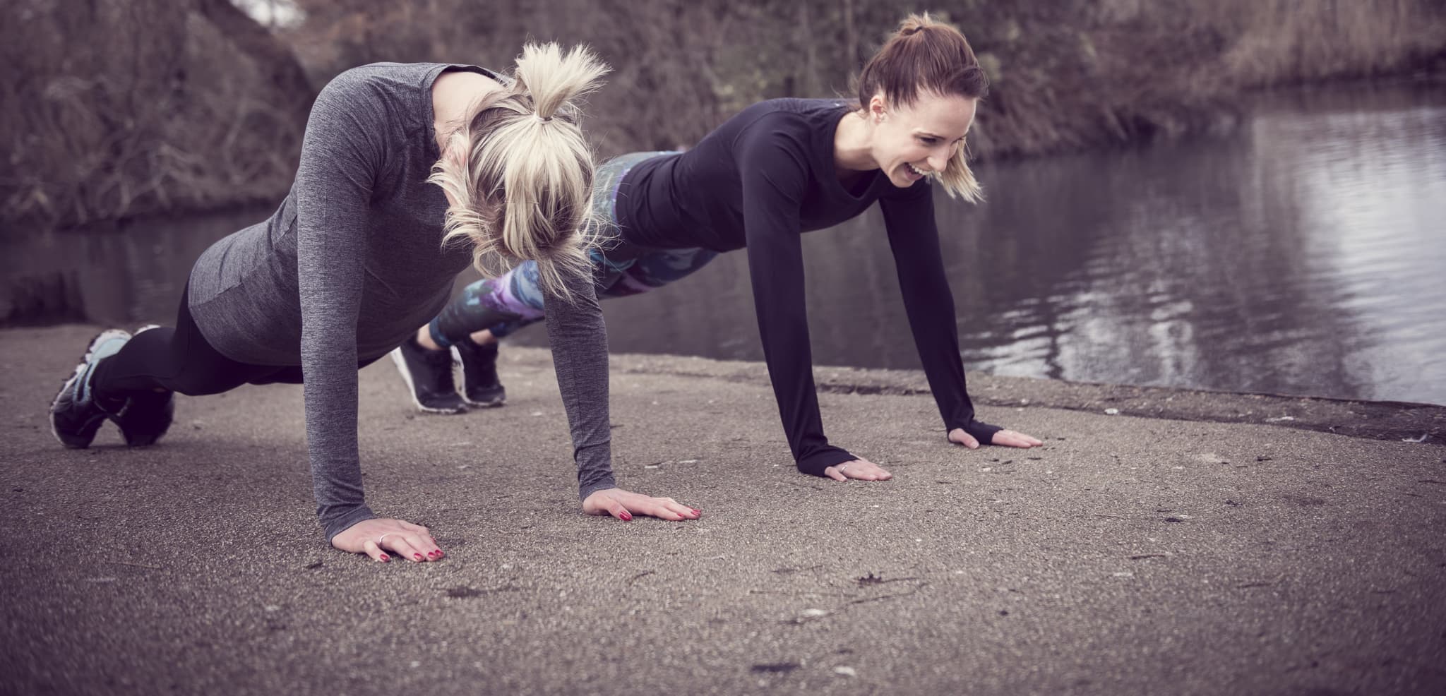 A photo of two women wearing gym clothes planking outdoors by a body of water.