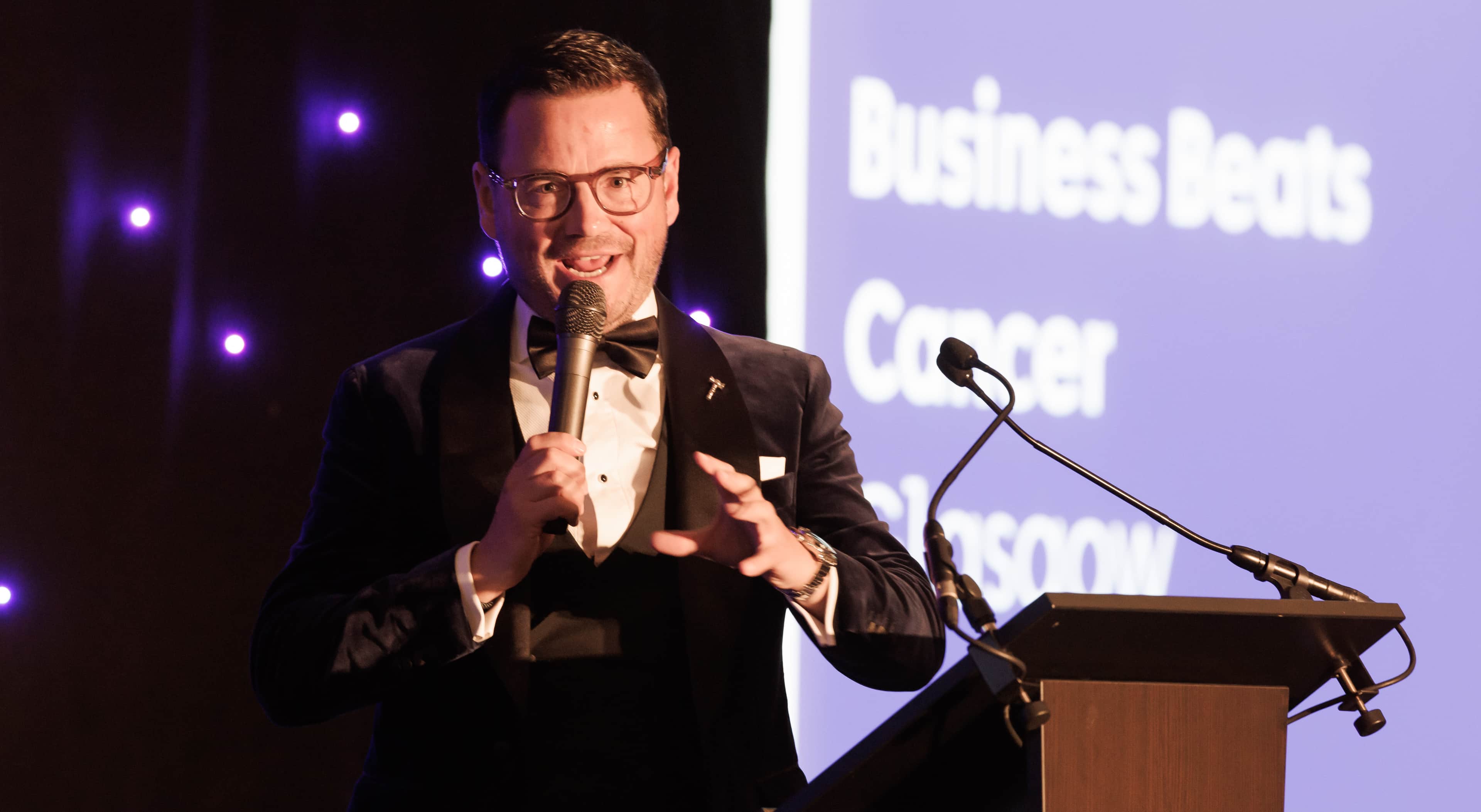 A man holding a microphone smiles to the camera, he is on a stage with the words 'Business Beats Cancer' behind him.
