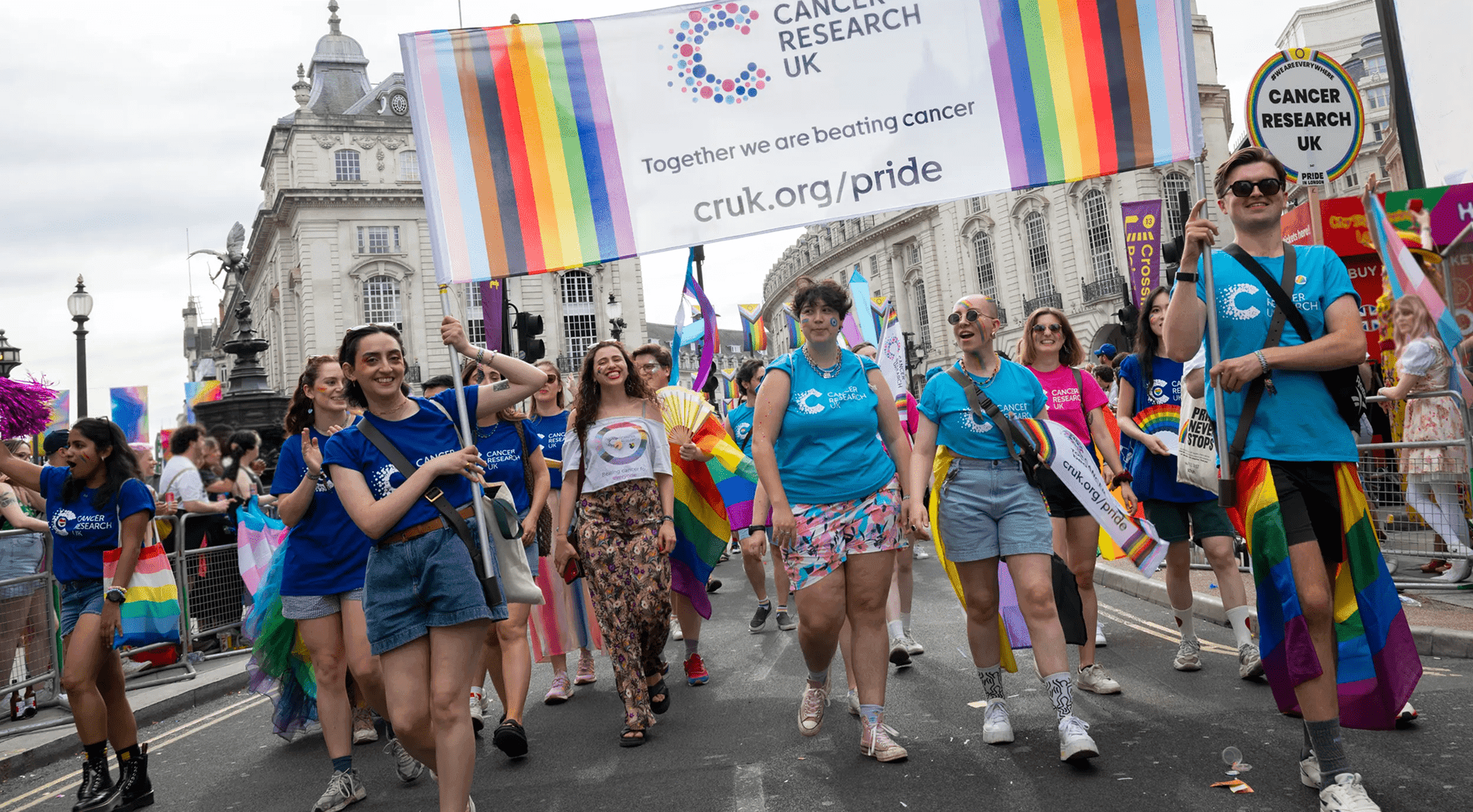 Group of smiling, happy people walking in a Pride parade and carrying a Cancer Research UK banner.