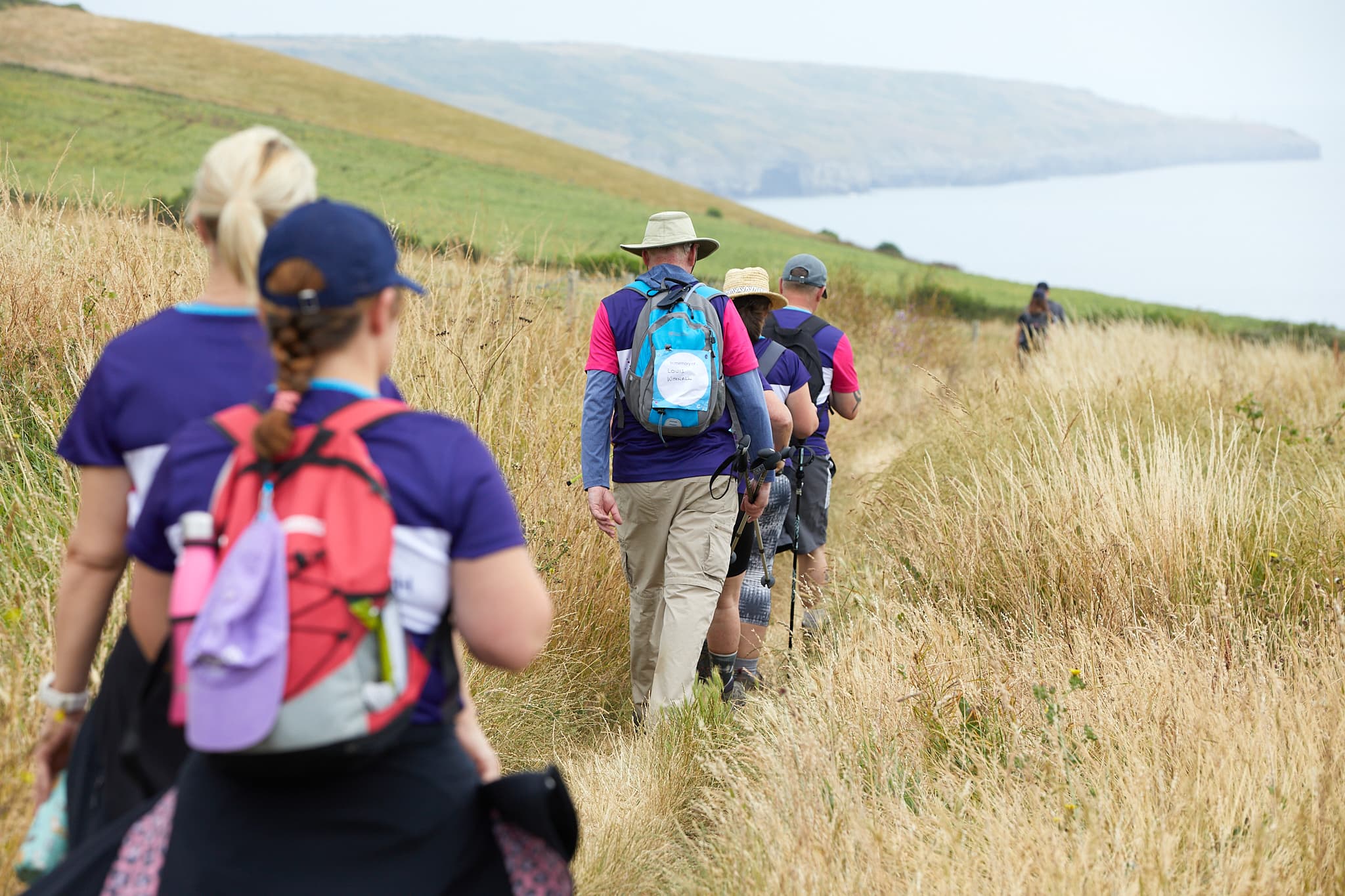 A group of hikers at a Cancer Research UK Big Hike event walking along a coastline with their backs towards the camera.