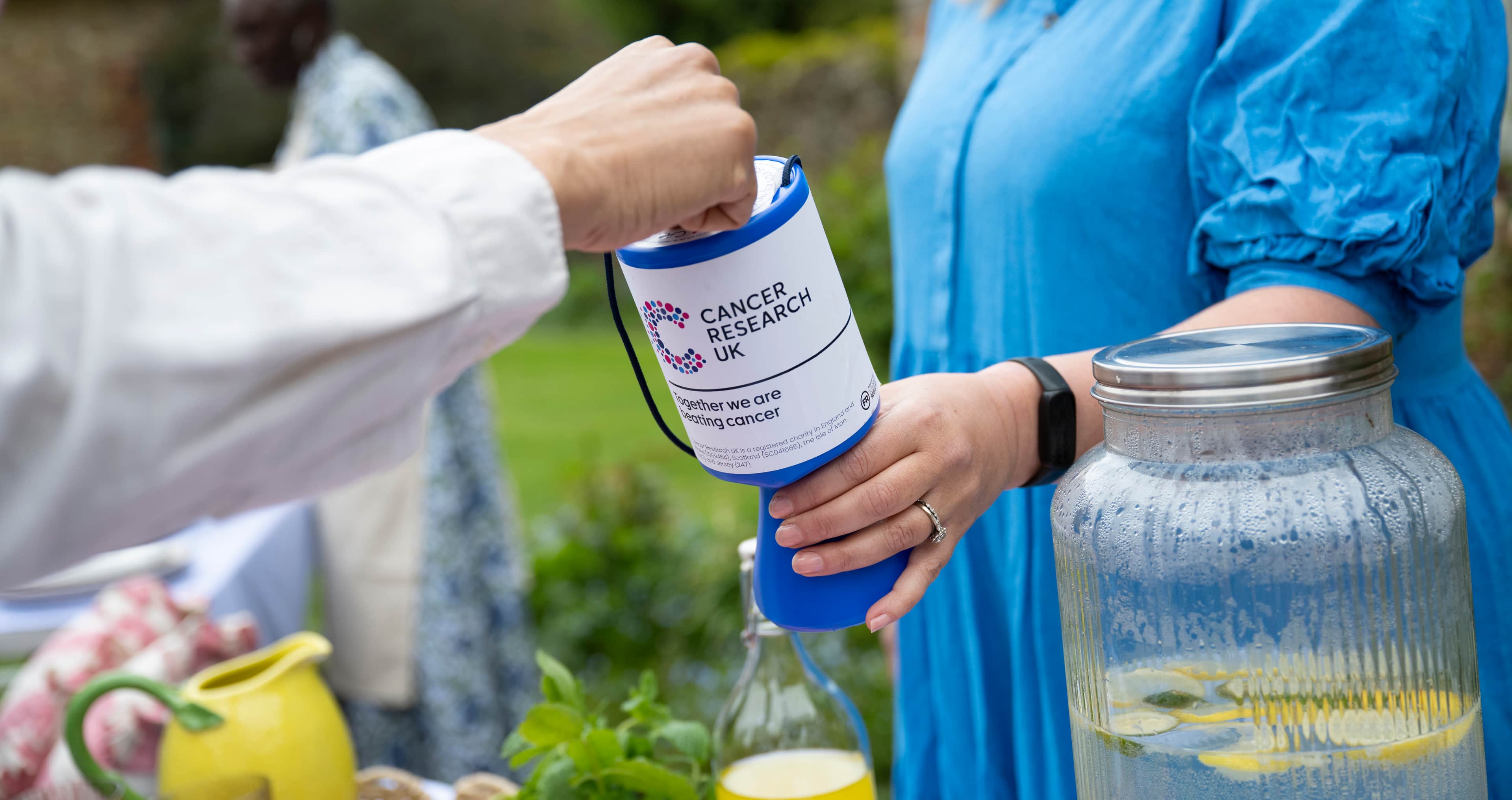 Person donating to Cancer Research UK tin at outdoor event, with lemonade jar visible in foreground.
