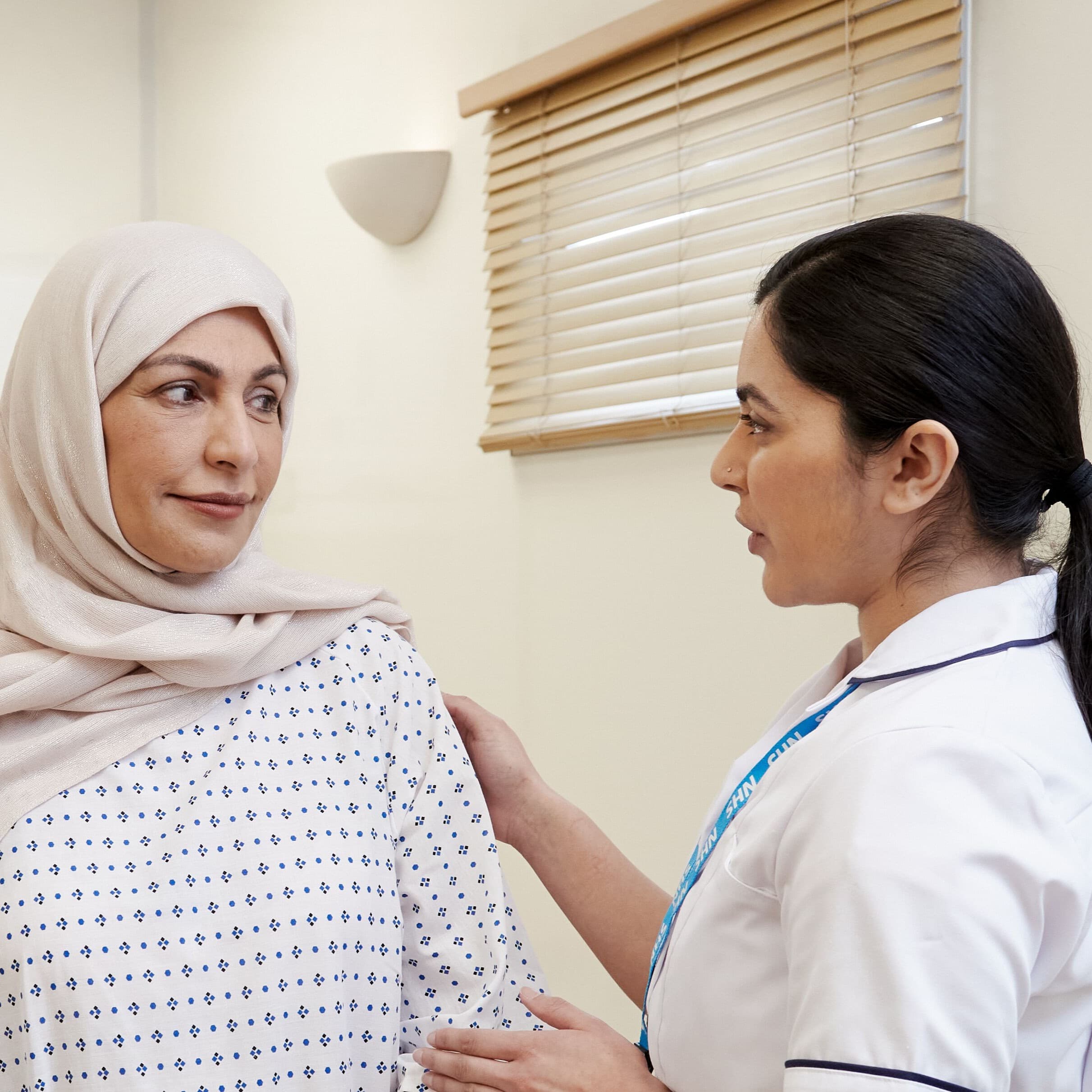 This photo shows a mammographer with a patient, wearing a hospital gown, before a mammogram.