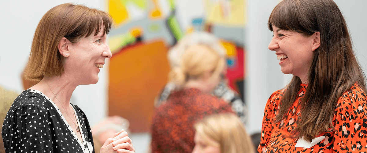 Two women standing in a room facing each other smiling.