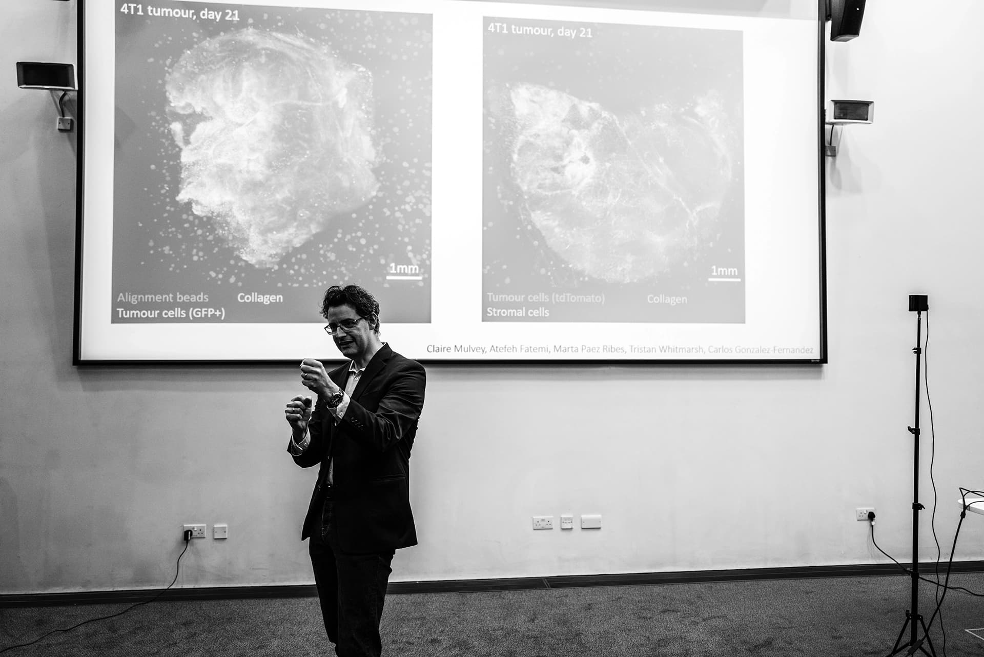 A male researcher in a suit, giving a lecture in front of a screen showing images of tumour scans.