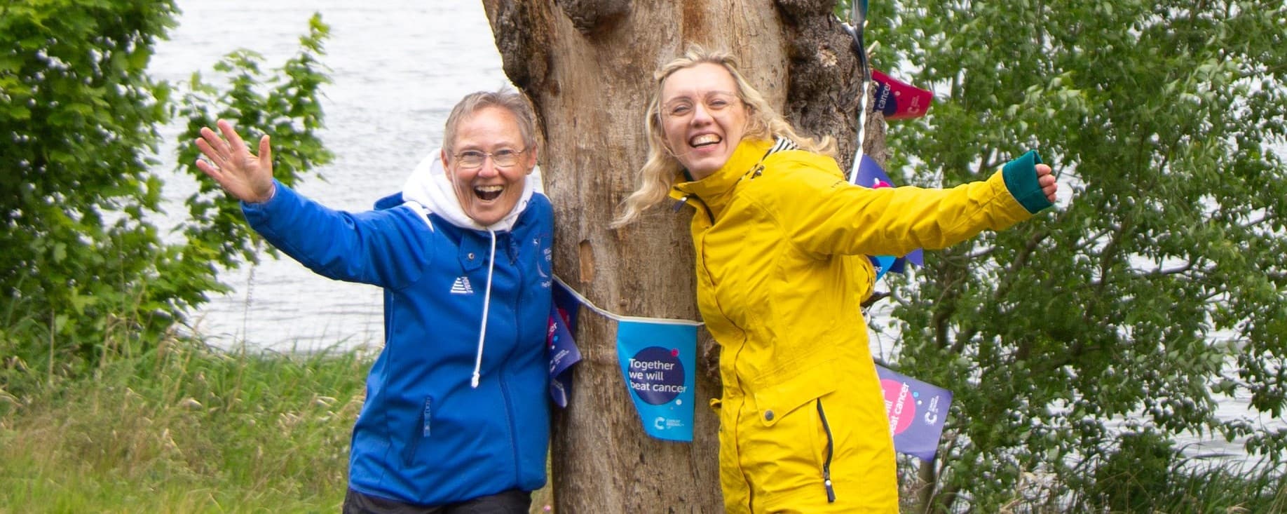 2 Relay for Life Peterhead Committee members posing with a tree next to a lake.