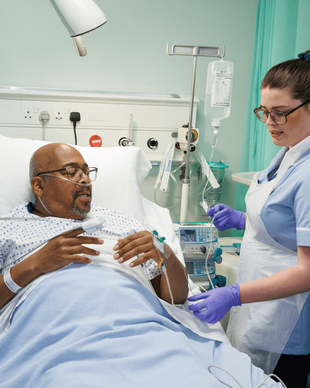 Patient in hospital bed with IV drip, attended by healthcare worker; medical equipment in background.