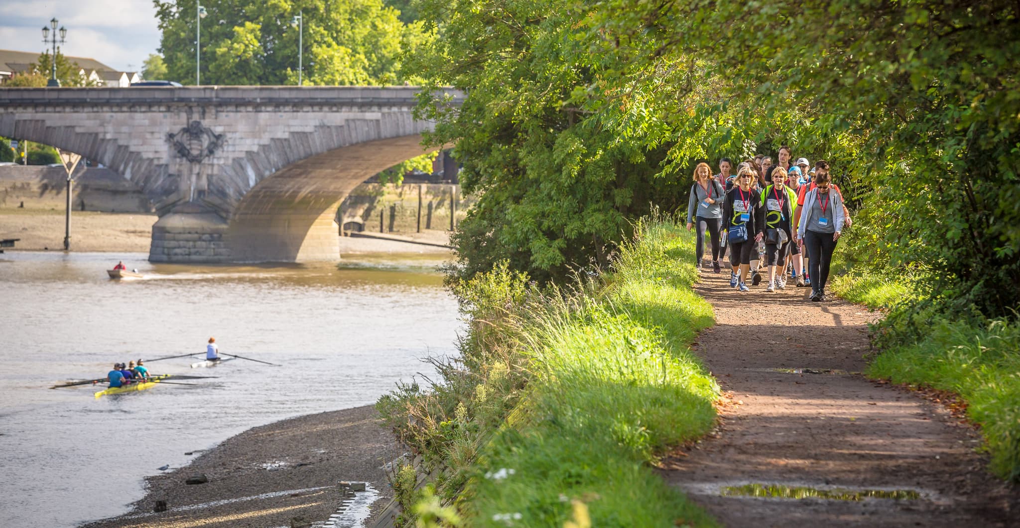 Group of walkers on the Thames Path next to the river with rowers in a boat about to go under a bridge.