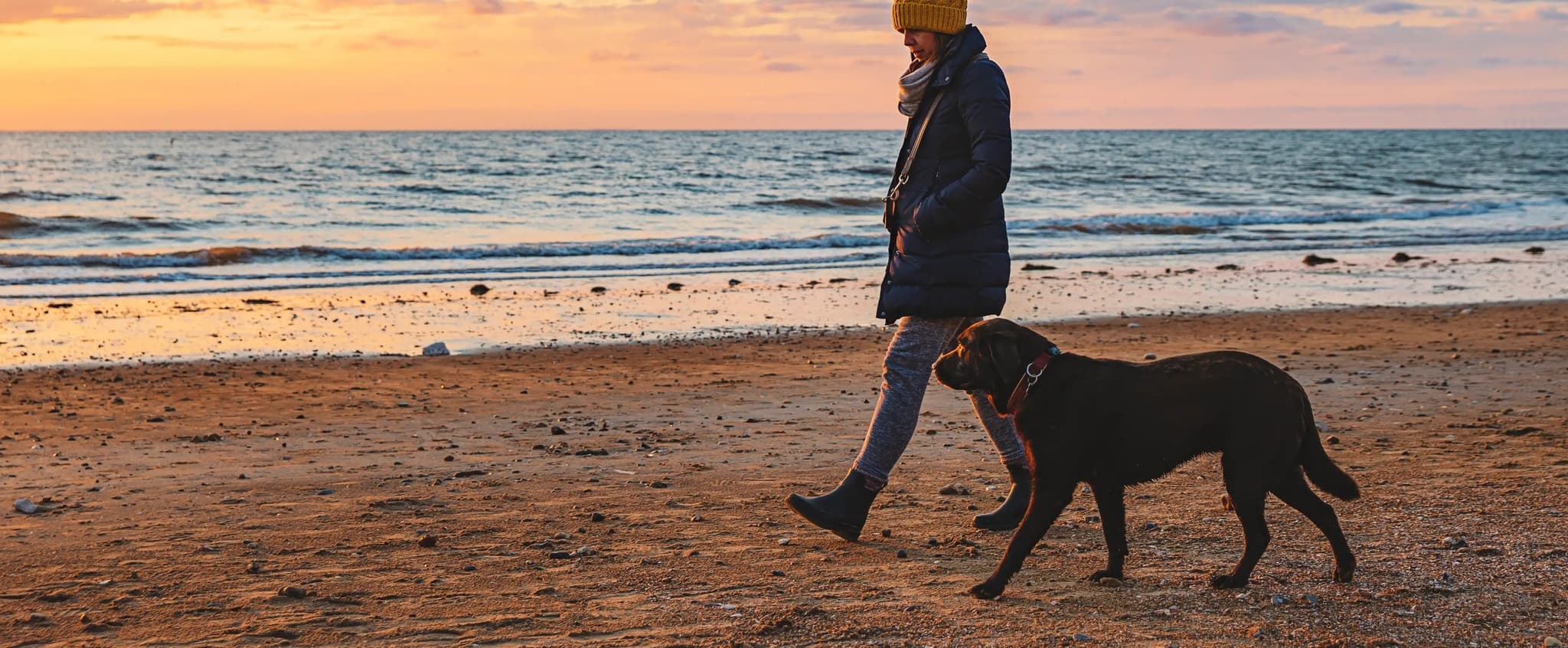 A lady in a yellow bobble hat walks with a chocolate Labrador on a beach at sunset.