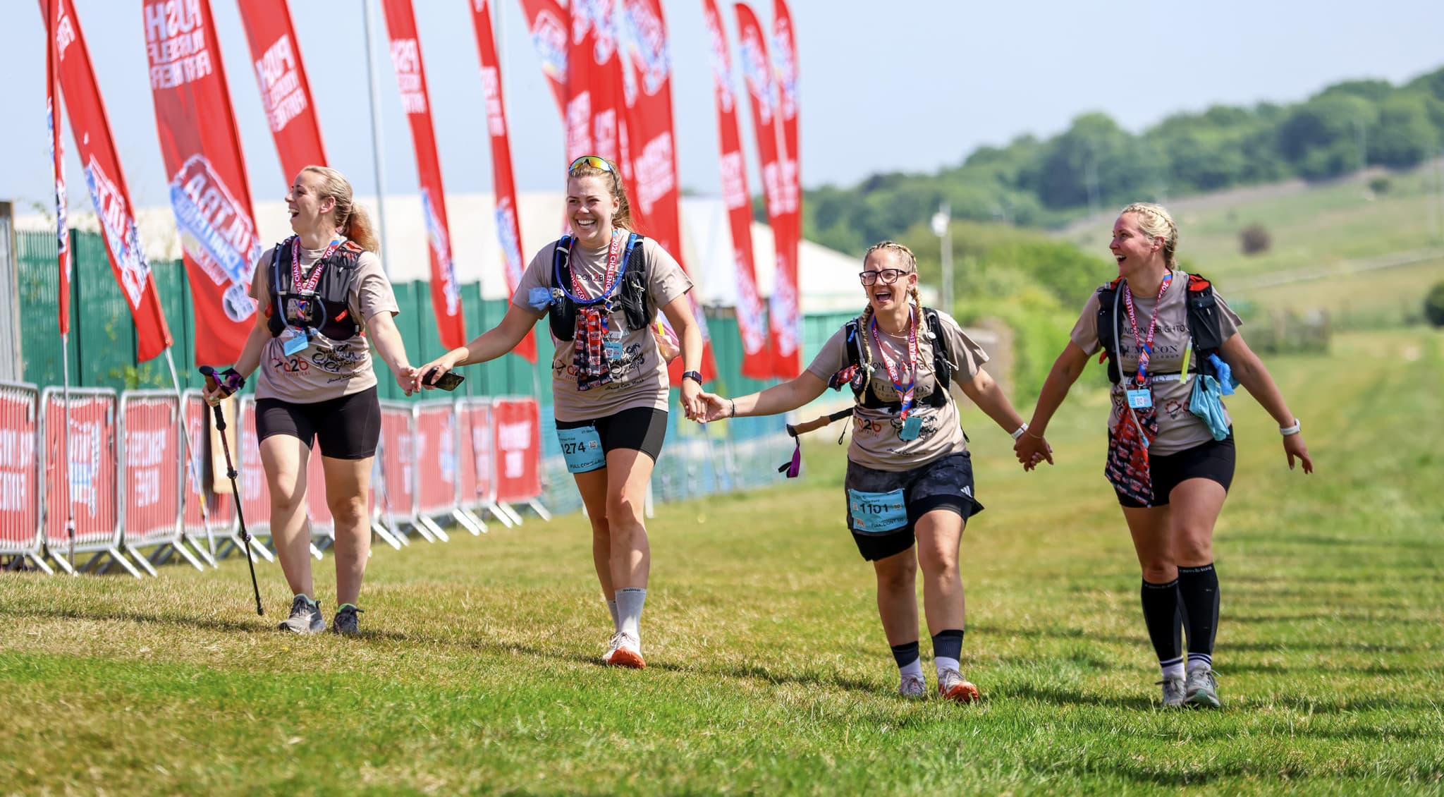 4 female participants holding hands walking through a field towards a finish line with flags to the side of them.
