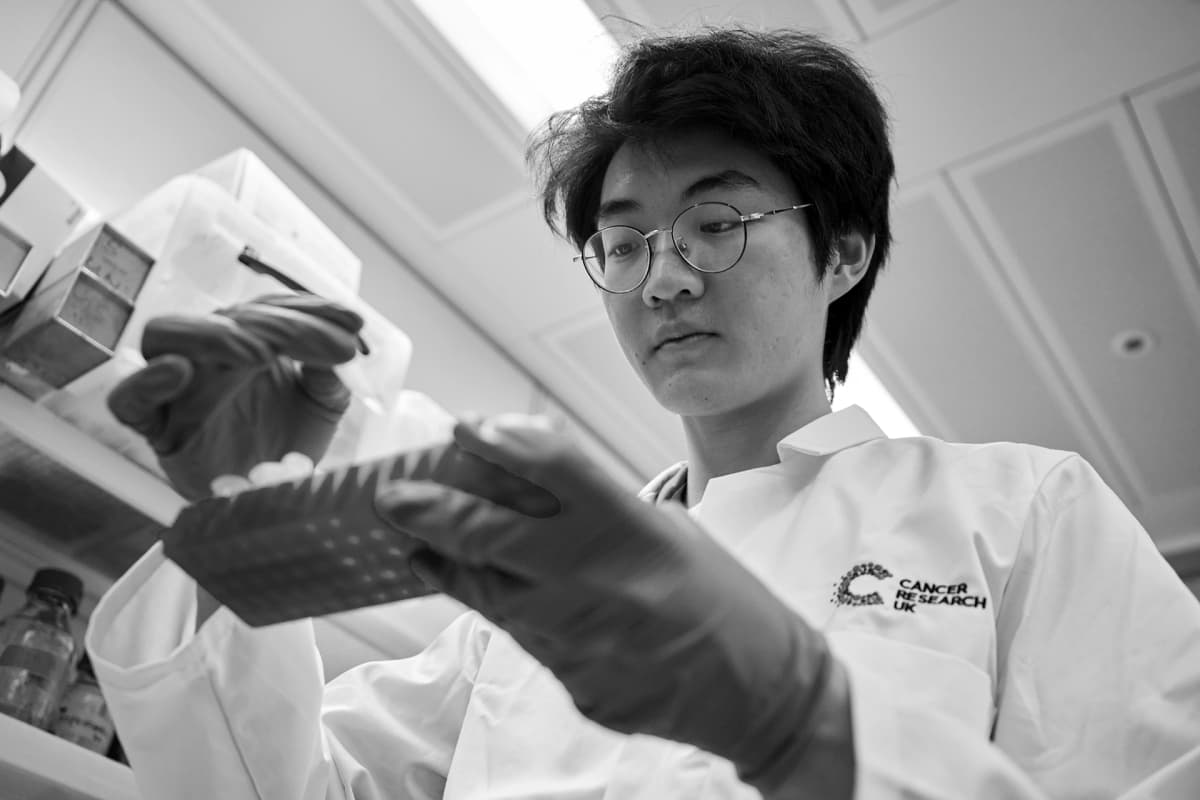 A black and white photograph of a male researcher wearing gloves in a laboratory, holding test tubes.