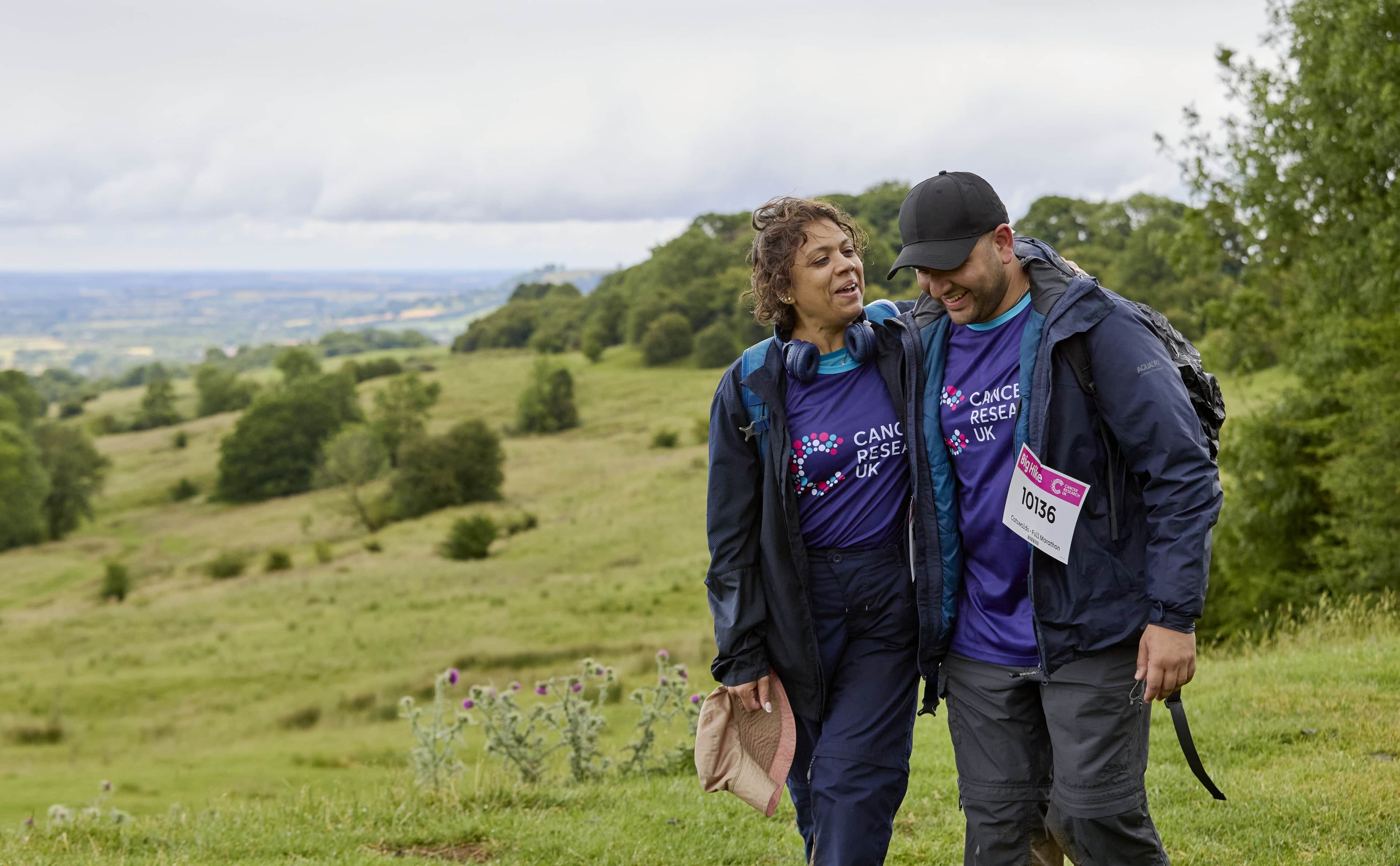 Two Big Hike participants, wearing Cancer Research UK shirts, stand close together on a grassy hill overlooking a valley.