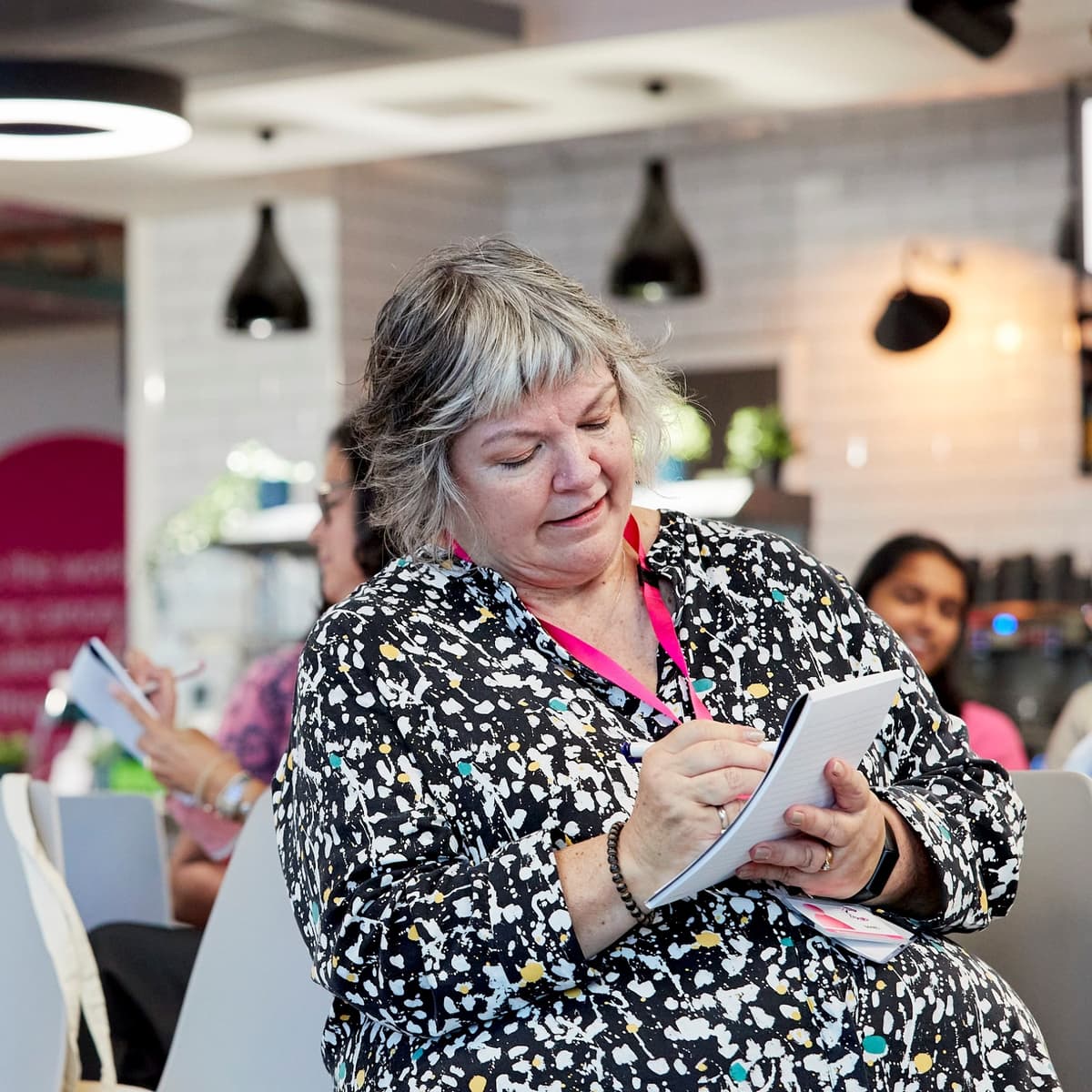 An image of a woman sat in a room that appears to be set up for an event. She is writing in a notebook.
