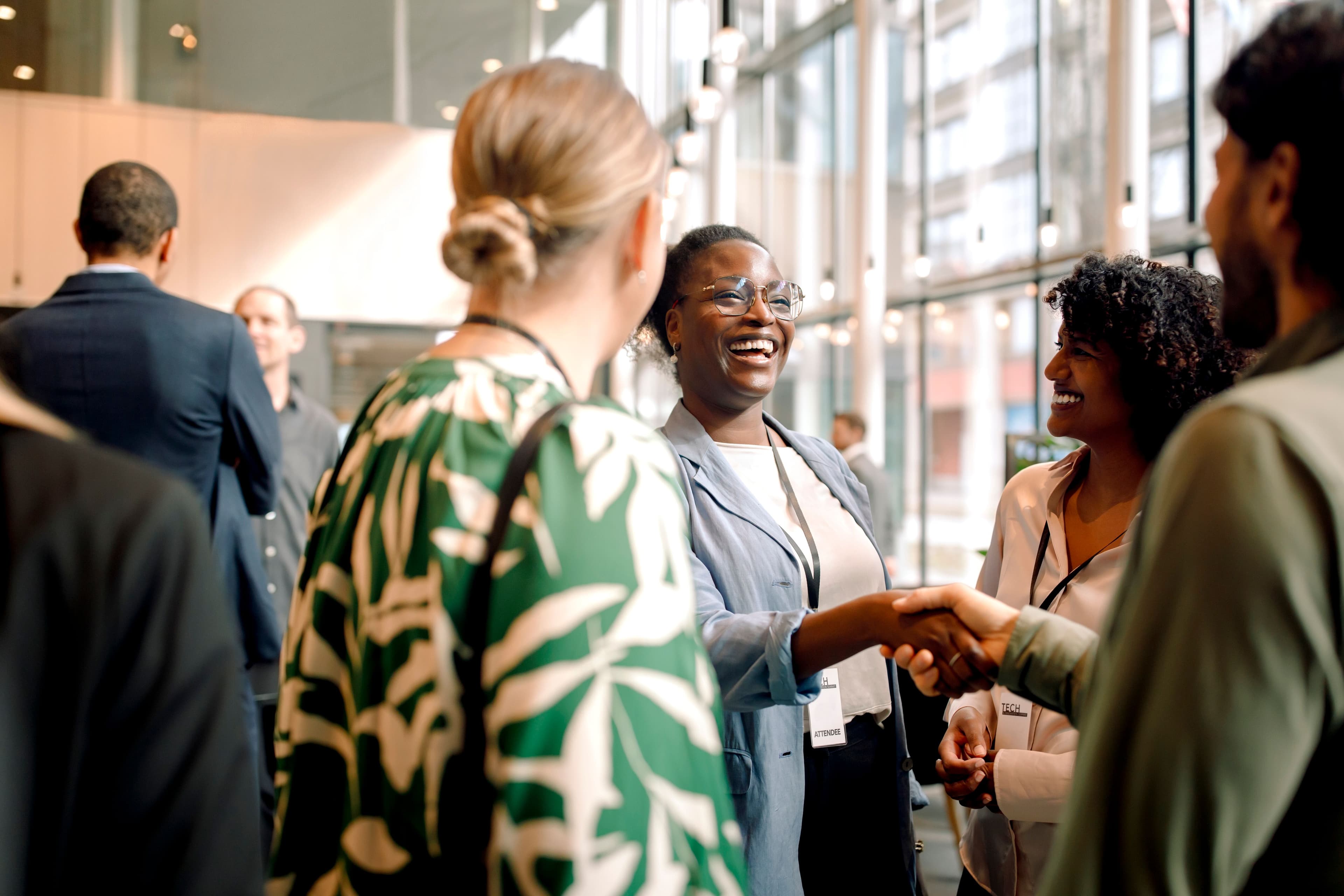 A smartly dressed, smiling woman shakes hands with a smartly dressed man at a gathering surrounded by smiling people.