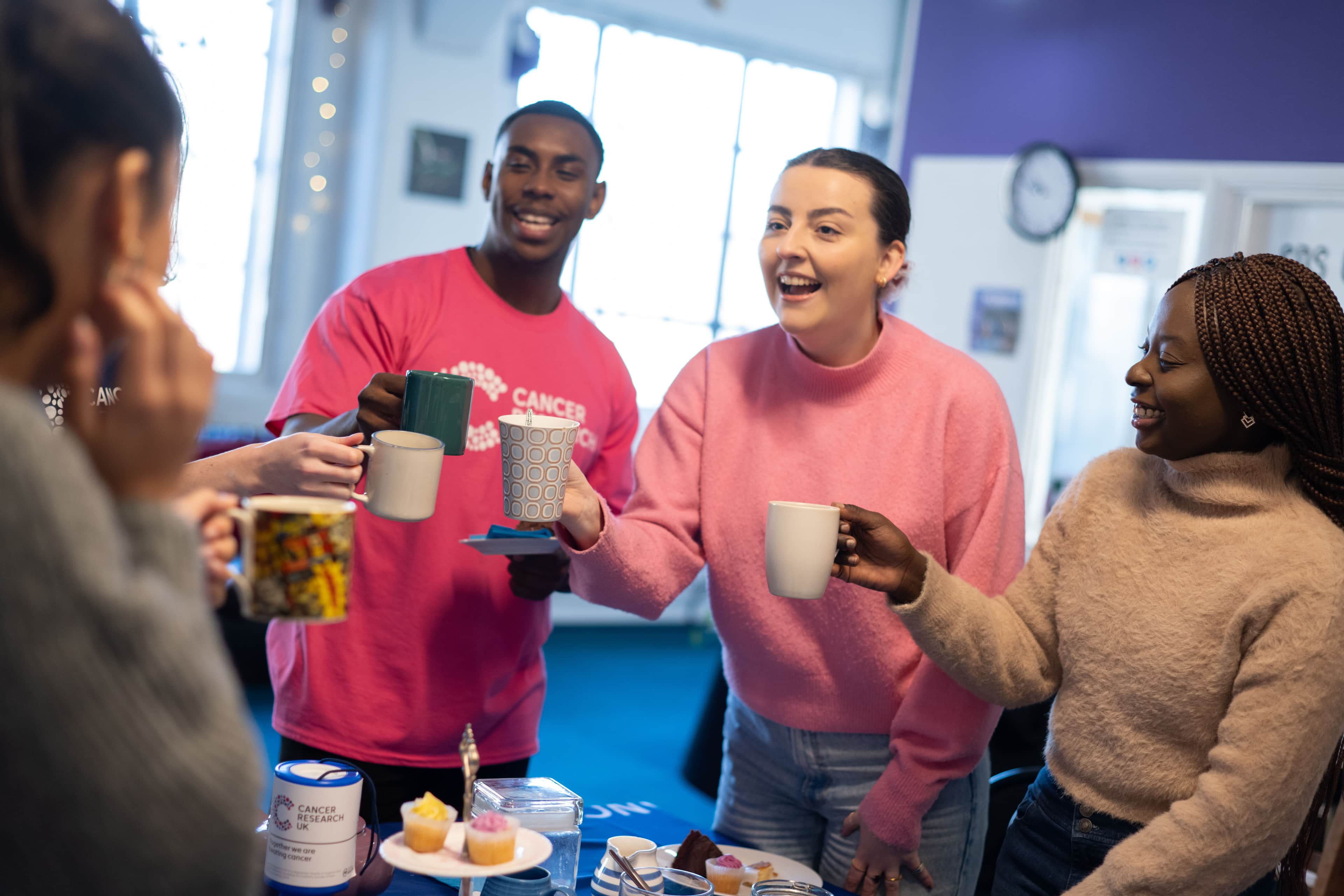 Cancer Research UK volunteers gather around a table with mugs and cake.