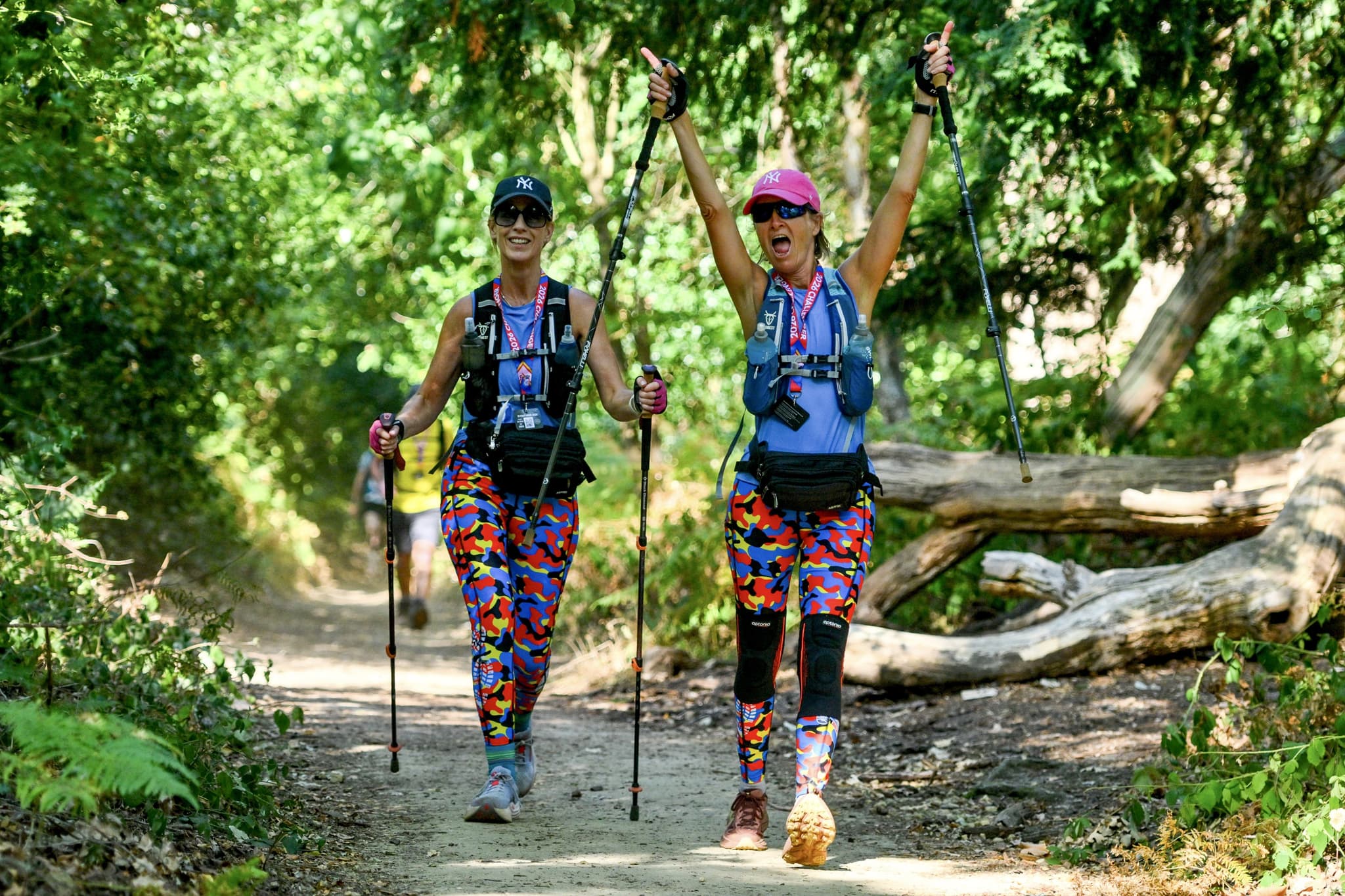 Two women walking through the woods with smiles on their faces & one with her arms raised in the air carrying walking poles.
