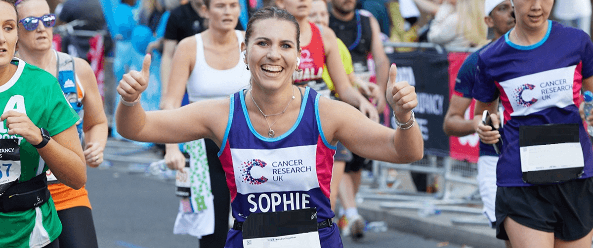 Female runner wearing a Cancer Research UK runner, smiling at the camera and giving thumbs-up as she takes part in a running event, surrounded by other runners.