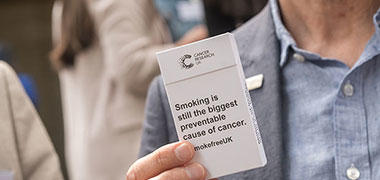 A photograph of someone holding a cigarette packet that reads 'Smoking is still the biggest preventable cause of cancer'.