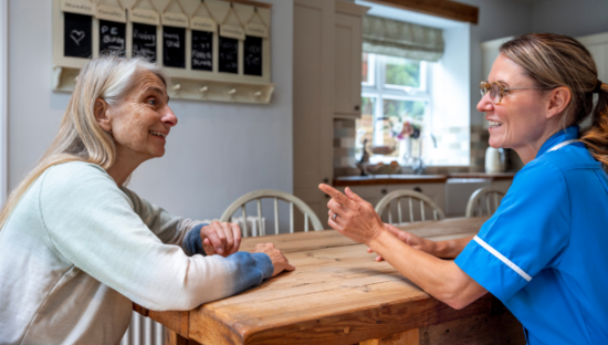 Medical professional talks to an elderly woman. They are sat at a table.