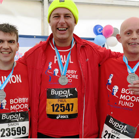 3 men wearing red, Bobby Moore Fund t-shirts, holding medals and smiling