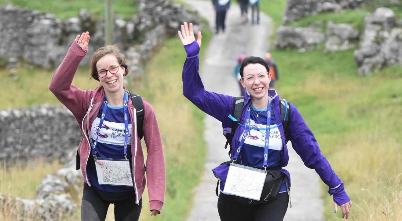 Big Hike participants walking along a mountainous path waving while wearing Cancer Research UK branded clothes.