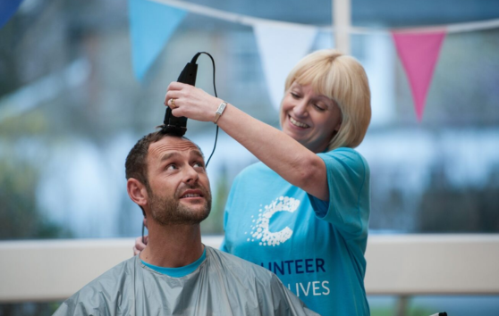 A woman wearing a Cancer Research UK top and shaving a man's head.