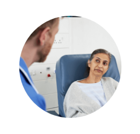 Donate monthly to help save more lives A female cancer patient sat in a chair in hospital talking to a nurse
