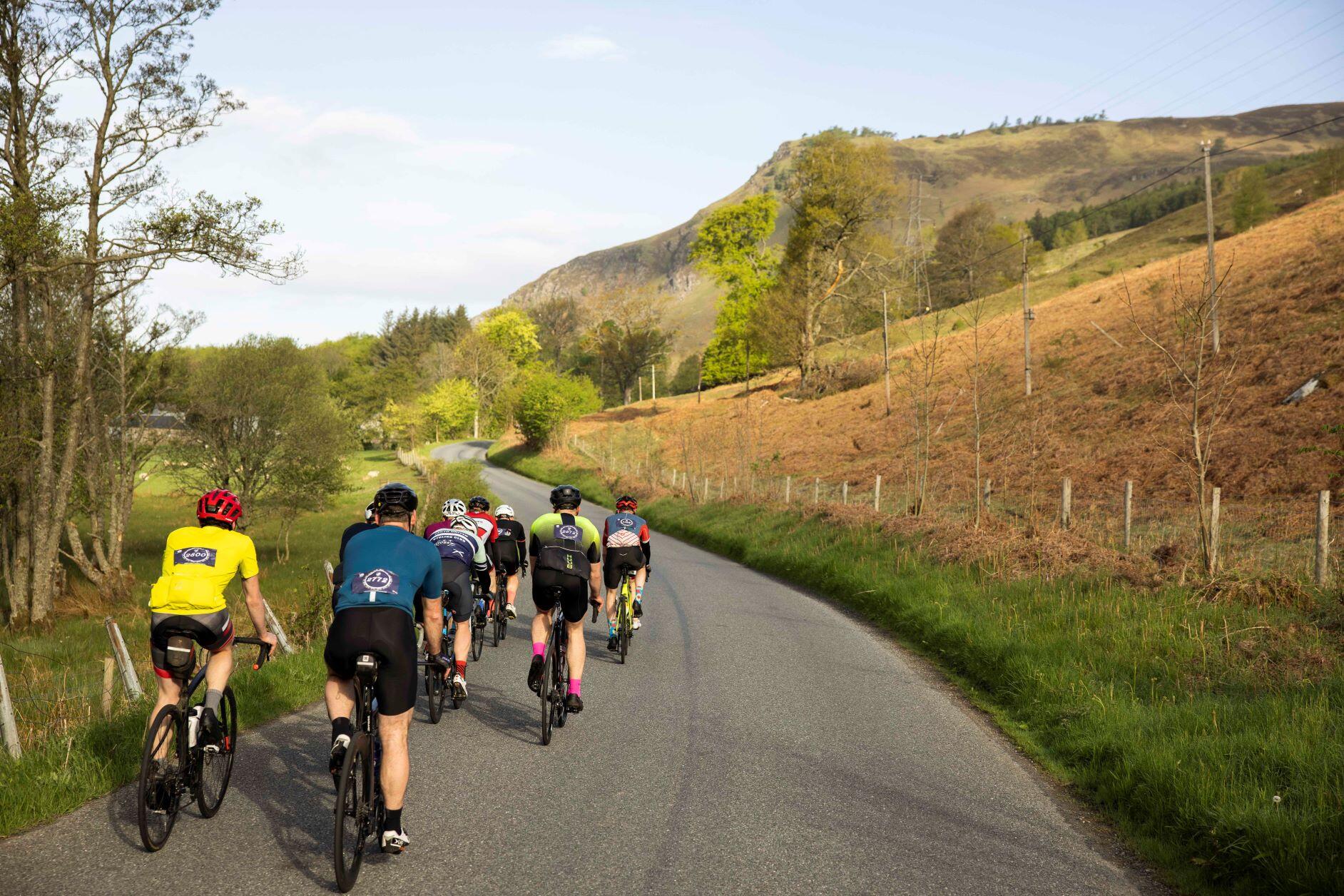 A group of cyclists (photographed from behind), cycling a country road with a backdrop of the Scottish highlands