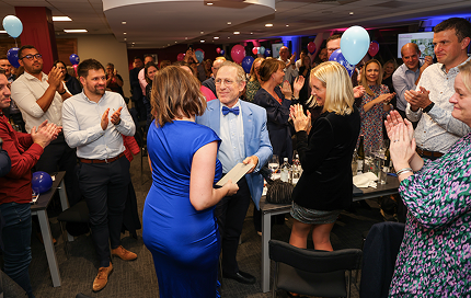 A man is giving a woman an envelope and group of people are clapping and celebrating at an event, surrounded by balloons
